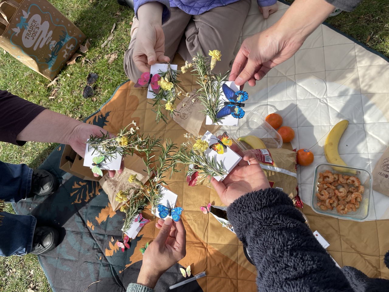 Overhead view of several hands over a picnic blanket, each holding a handmade card with a paper butterfly and a sprig of rosemary.